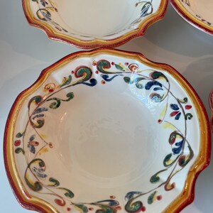 May include: A set of white ceramic bowls with a scalloped edge. Each bowl features a decorative border with red, yellow, and green accents. The interior of the bowls is plain white, and the design suggests a rustic or Mediterranean style.