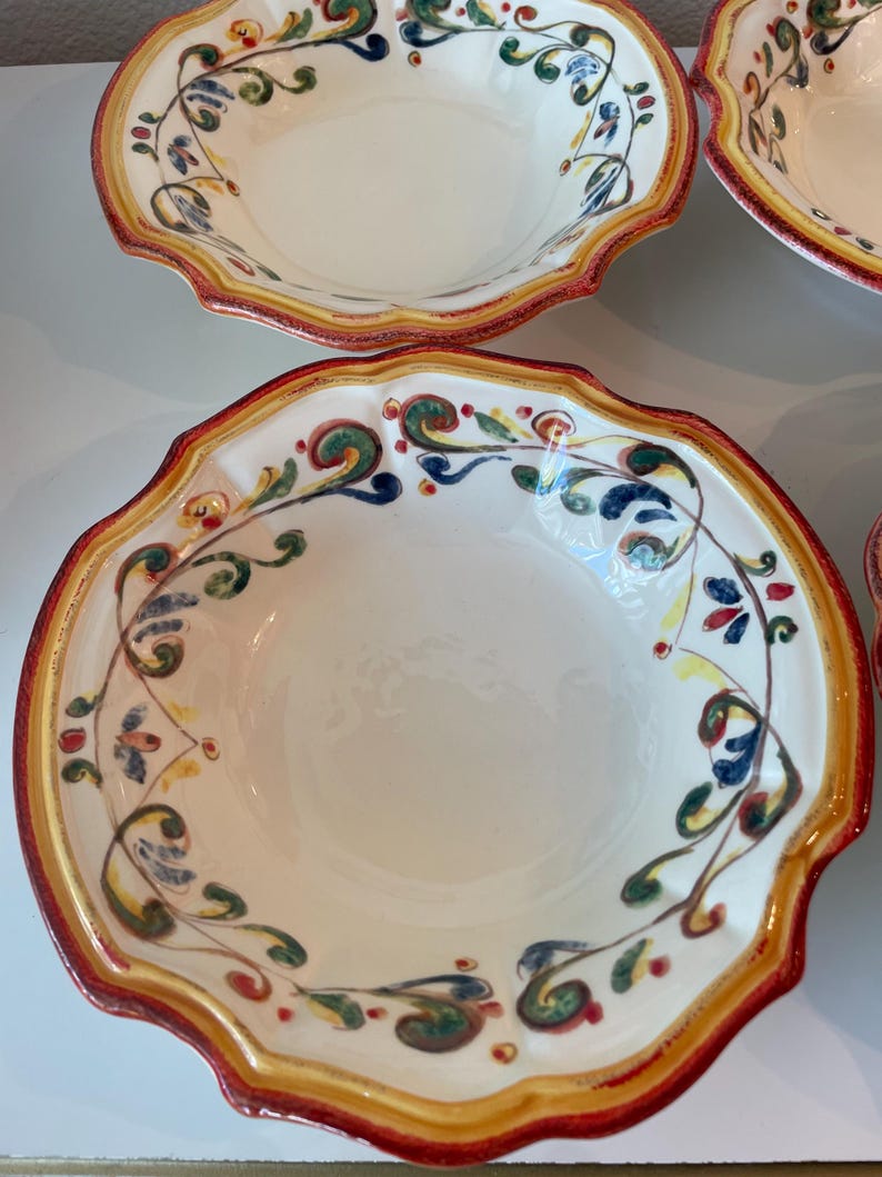 May include: A set of white ceramic bowls with a scalloped edge. Each bowl features a decorative border with red, yellow, and green accents. The interior of the bowls is plain white, and the design suggests a rustic or Mediterranean style.