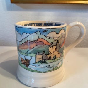 May include: White ceramic mug with a handle, featuring a colorful illustration of a Scottish Highlands landscape. The design includes a castle, mountains, a lake, and a Highland cow. The text "SCOTTISH HIGHLANDS" is printed on the rim.