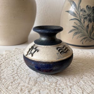 May include: A small, round ceramic vase featuring a dark blue glazed base and neck, with a contrasting cream-colored band around the center. The cream section is decorated with two distinct, dark, embossed symbols, one resembling a star and one wavy lines.