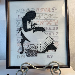 以下が含まれることがあります： 額縁に入ったクロスステッチのアートワーク。裁縫をする女性のシルエットが描かれ、背景にはアルファベットと数字があります。作品は濃い木製のフレームに入っており、「1998」の年号とイニシャルが含まれています。装飾的な金属製のスタンドに展示されています。