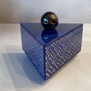 Puede incluir: Una caja triangular azul con un patrón plateado y una esfera gris oscuro brillante en la parte superior. La caja tiene un acabado brillante y un diseño geométrico, adecuada para guardar objetos pequeños.