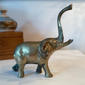 May include: A brass elephant figurine with an elongated, curved trunk and raised tusks. The elephant has detailed features, including wrinkles and textured skin. The figurine is standing on four legs and has a slightly aged appearance, suggesting it is vintage.