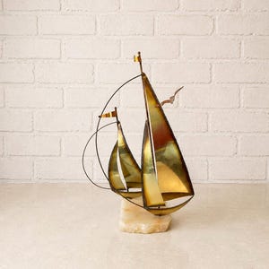 Puede incluir: Una escultura de metal de un velero de dos mástiles con velas de latón pulido y bordes oscuros. La estructura incluye un aparejo de alambre fino y curvado y pequeñas banderas en la parte superior de cada mástil sobre una base de piedra.