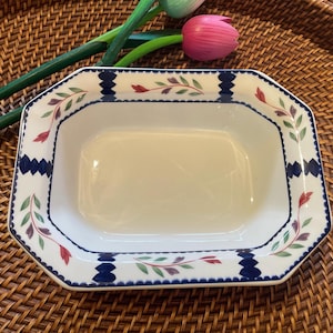 May include: A rectangular, off-white serving dish with a decorative border. The border features a repeating pattern of blue, green, and red floral elements. The dish is set on a woven brown surface, with artificial flowers in the background.