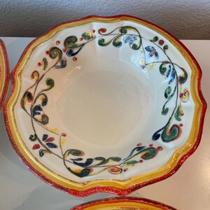 May include: Off-white ceramic bowl with a scalloped edge and a colorful floral design. The rim is bordered with yellow and red paint. The bowl's interior is plain, and the floral design includes green, blue, and red accents.