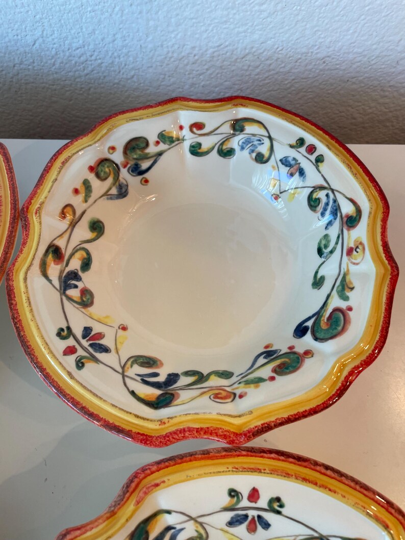 May include: Off-white ceramic bowl with a scalloped edge and a colorful floral design. The rim is bordered with yellow and red paint. The bowl's interior is plain, and the floral design includes green, blue, and red accents.