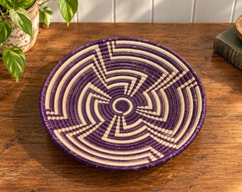 May include: A round, shallow woven basket featuring a geometric pattern in deep purple and cream. The design consists of concentric circles and angular lines radiating from a central spiral, creating a structured, repeating visual motif.
