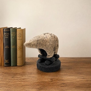 Op de afbeelding: Een keramieken sculptuur van een beer met een gebarsten, gebroken witte en grijs gespikkelde afwerking. De beer staat op een donkere, getextureerde ronde voet. Links van de beer staan drie boeken met bruine, groene en beige ruggen.