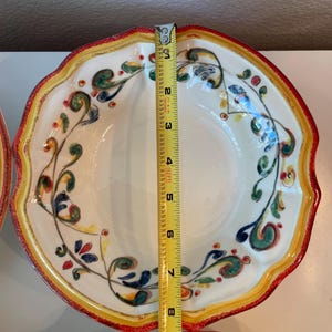 May include: White ceramic bowl with a scalloped edge, measuring approximately 9 inches in diameter. The bowl features a decorative floral design in green, blue, and red, with a yellow and red border. The bowl is part of a set.