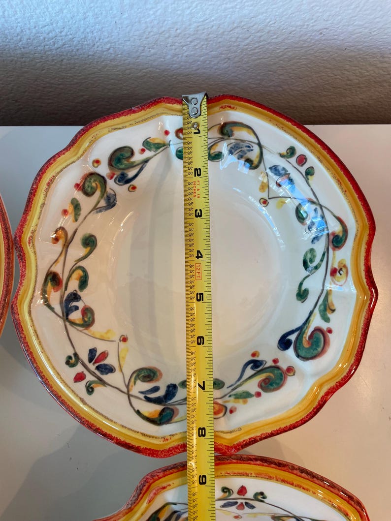 May include: White ceramic bowl with a scalloped edge, measuring approximately 9 inches in diameter. The bowl features a decorative floral design in green, blue, and red, with a yellow and red border. The bowl is part of a set.