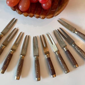 May include: A collection of vintage fruit forks and knives with wooden handles and brass accents. The forks and knives are arranged around a wooden bowl filled with red grapes, creating a classic and elegant presentation.