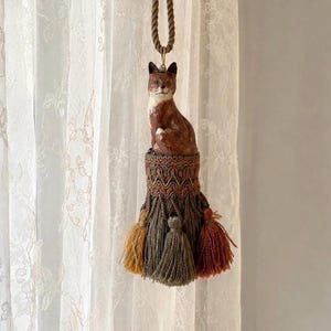 May include: A decorative hanging tassel featuring a sculpted brown and white fox figure perched atop a woven cylindrical base. The base is adorned with intricate patterns and finished with multiple hanging yarn tassels in mustard, olive, and rust colors.