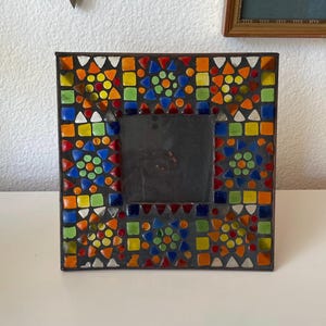 May include: A square picture frame featuring a mosaic border composed of small, multi-colored glass tiles in shades of red, orange, yellow, green, and blue. The tiles are arranged in geometric patterns around a central dark, square opening.