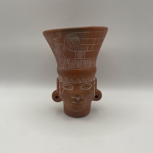 May include: A terracotta-colored ceramic vessel shaped like a human head with a flared rim. The surface features intricate white etched line patterns, including geometric shapes, circular motifs, and stylized facial features on the front.
