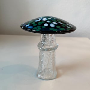 Pode incluir: Um cogumelo decorativo de vidro com um chapéu azul escuro adornado com pontos verdes, brancos e azuis. A haste é transparente e texturizada. Esta peça de arte única adiciona um toque caprichoso a qualquer decoração.