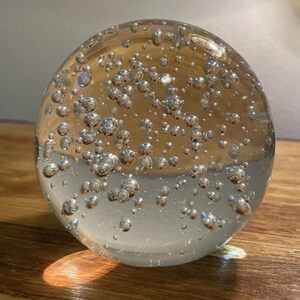 Puede incluir: Una esfera de vidrio transparente llena de numerosas burbujas plateadas. La esfera descansa sobre una superficie de madera, reflejando la luz y creando sombras. Las burbujas varían en tamaño, agregando interés visual al objeto decorativo.