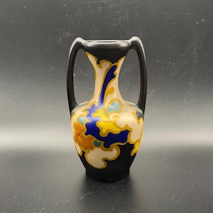 Peut inclure: Petit vase décoratif avec une base et des anses noires. Le vase présente un corps crème avec un motif floral coloré dans les tons de bleu, jaune et orange. Les anses et le bord supérieur sont noirs.