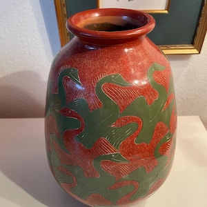 May include: A red ceramic vase with a green swan design. The vase has a wide body and a narrow neck. The swans are arranged in a repeating pattern around the vase. The vase is likely used for decorative purposes.