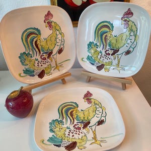 Puede incluir: Tres platos cuadrados de cerámica con esquinas redondeadas, cada uno con un diseño de gallo pintado a mano. Los gallos están representados en amarillo, verde, azul y rojo. Los platos se exhiben sobre pequeños caballetes de madera, con una figura de manzana roja en primer plano.