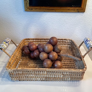 Puede incluir: Una cesta rectangular de mimbre tejido con asas decorativas. Las asas tienen un marco de metal y extremos de cerámica con un diseño floral azul, amarillo y blanco. La cesta contiene un grupo de esferas de madera marrón.