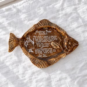May include: A brown glazed ceramic fish-shaped serving platter featuring a white printed recipe for Gustavus Baked Halibut, including a chef illustration and the text Tom's Pots, Juneau, Alaska, with instructions for oven temperatures in degrees Celsius.