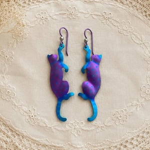 May include: A pair of dangle earrings featuring a cat silhouette design. The cats are colored in a gradient of purple and blue with small light blue polka dots. Each earring hangs from a purple curved ear wire with a small silver-toned bead.