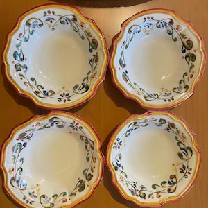 May include: Four white ceramic bowls with scalloped edges. Each bowl features a decorative floral pattern in green, blue, and red, with a yellow and red border. The bowls are arranged on a wooden surface.