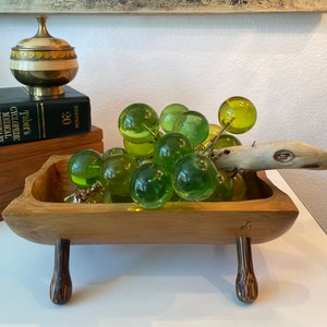 Puede incluir: Un cuenco de madera con un arreglo decorativo de uvas de cristal verde y un trozo de madera flotante. El cuenco tiene una forma única y está sostenido por dos patas cortas de color oscuro. Un pequeño recipiente de latón y un diccionario médico están en el fondo.