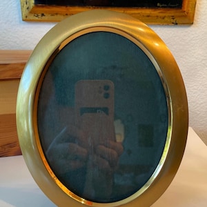 May include: Oval, gold-toned picture frame with a dark green interior. The frame has a simple, elegant design and rests on three small, rounded feet. A painting with a gold frame is visible in the background.