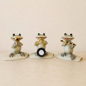 May include: Three light green ceramic frog figurines arranged in a row. The left frog plays a gold trumpet, the center frog plays a drum with yellow sticks, and the right frog plays a yellow flute. Each frog sits on a small, curved white base.