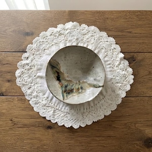 May include: A round, off-white ceramic bowl with a speckled texture and a prominent, abstract dark blue and brown glaze pattern across the center, resting on a circular white lace doily with a scalloped edge and intricate floral embroidery.