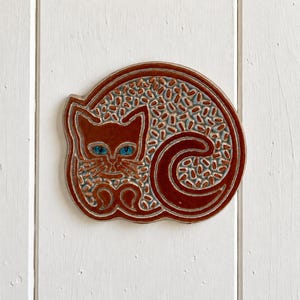 Könnte beinhalten: Eine kreisförmige Wandplatte aus Keramik mit einer stilisierten, zusammengerollten Katze. Die Katze ist in einer tief rotbraunen Glasur mit leuchtend blauen Augen gehalten. Der Hintergrund der Platte ist mit einem strukturierten Muster aus kleinen hellblauen Strichen gefüllt.