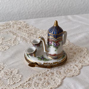 May include: A miniature porcelain tea set featuring a white teapot with blue vertical stripes, gold trim, and floral accents. The set includes two small cups and a tiny spoon, all arranged on a matching oval tray with painted green and blue floral details.
