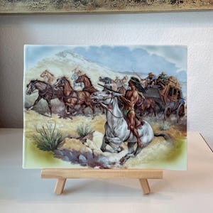 Può includere: Piastrella in ceramica con un'illustrazione a tema western di una diligenza inseguita da cavalieri nativi americani a cavallo. L'opera d'arte raffigura una scena dinamica con cavalli, cavalieri e una diligenza sullo sfondo di un paesaggio desertico.