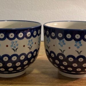 May include: Two ceramic mugs with a blue and white floral pattern. The mugs have a rounded shape with a handle and a wide rim. The design includes blue flowers and dark blue polka dots. The mugs are on a wooden surface.