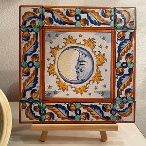 Puede incluir: Azulejo cerámico cuadrado con diseño celestial. La imagen central representa una luna creciente con rostro, rodeada de un sol y estrellas. El borde tiene un patrón decorativo en azul, naranja y verde. El azulejo se exhibe sobre un pequeño caballete de madera.