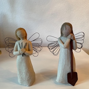 May include: Two angel figurines with wire wings. One angel holds a bundle of greenery, the other leans on a shovel. Both wear long, textured white dresses and have simple, neutral-toned faces. The figurines are on a white surface.