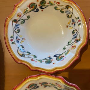 May include: White ceramic bowls with scalloped edges and colorful floral designs. The rims are bordered with red and yellow paint. The interior of the bowls is a creamy white color. The floral design includes green, blue, and red accents.
