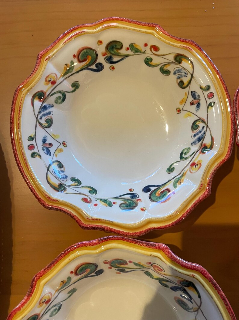 May include: White ceramic bowls with scalloped edges and colorful floral designs. The rims are bordered with red and yellow paint. The interior of the bowls is a creamy white color. The floral design includes green, blue, and red accents.