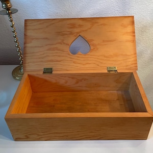 Könnte beinhalten: Offene Holzkiste mit herzförmigem Ausschnitt im Deckel. Die Kiste ist aus hellem Holz gefertigt und hat Messing Scharniere. Der Innenraum ist leer und die Kiste ist rechteckig.