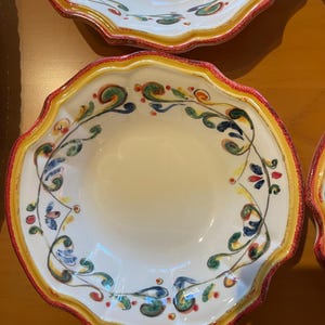 May include: A set of white ceramic bowls with scalloped edges. Each bowl features a decorative floral pattern in green, blue, and red, with a yellow and red border. The bowls are designed for serving food.