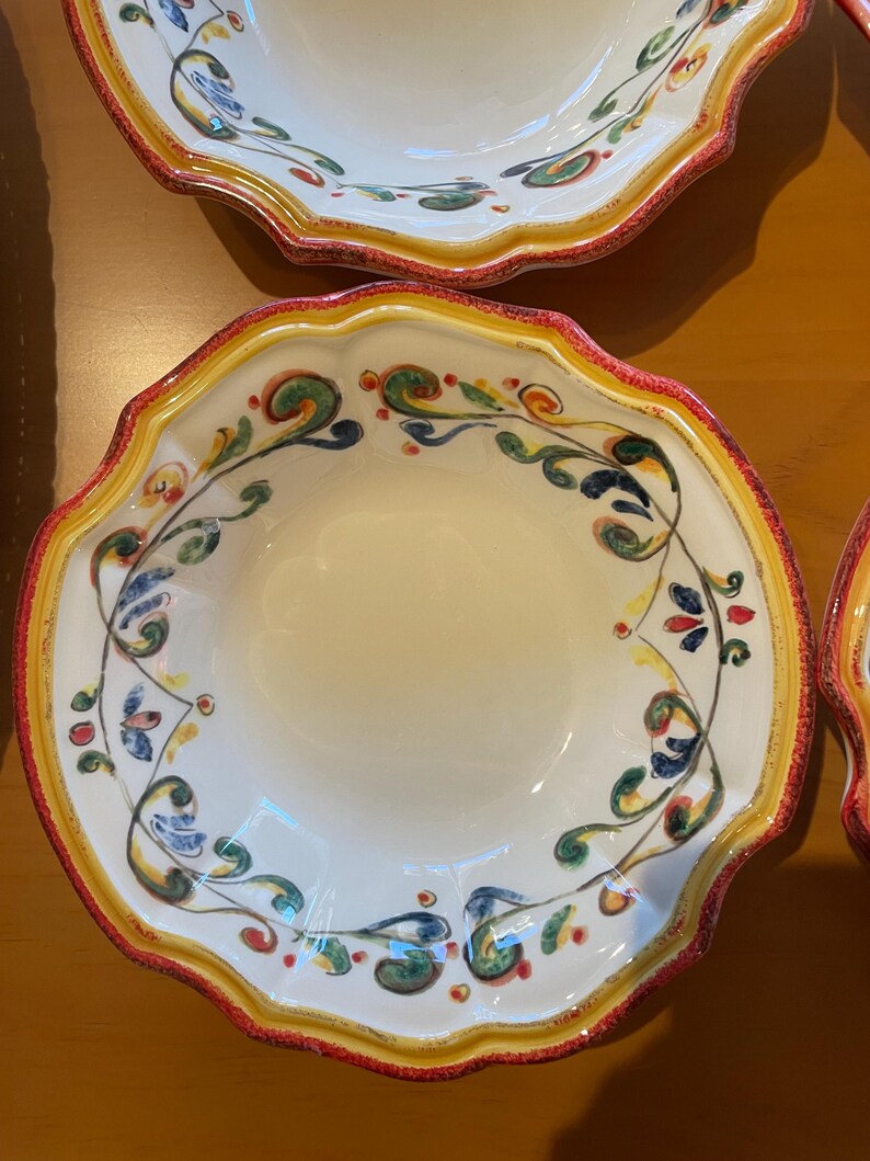 May include: A set of white ceramic bowls with scalloped edges. Each bowl features a decorative floral pattern in green, blue, and red, with a yellow and red border. The bowls are designed for serving food.