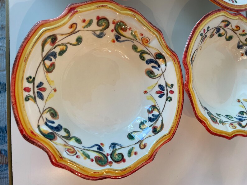 May include: White ceramic bowls with scalloped edges and colorful floral designs. The rims are trimmed with red, yellow, and orange paint. The bowls have a decorative pattern of green, blue, and red leaves and flowers.