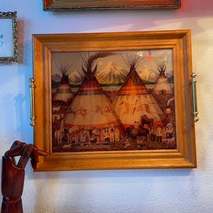 Może przedstawiać: Oprawiony wydruk przedstawiający scenę wioski rdzennych Amerykanów z tipi, postaciami i górami w tle. Wydruk jest w jasnobrązowej drewnianej ramie ze złotymi uchwytami po bokach.