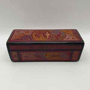May include: A rectangular black wooden box with a hinged lid, featuring intricate hand-painted designs in red, yellow, and purple. The lid and front panel display detailed illustrations of birds, floral patterns, and small animals with fine line work.