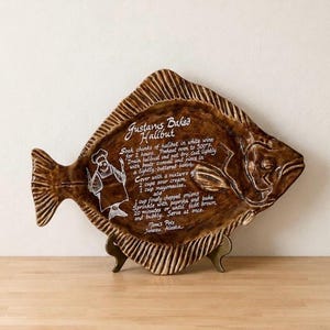 May include: A brown, fish-shaped ceramic platter featuring a white graphic of a chef and the recipe for Gustavus Baked Halibut. The text includes instructions for preparing halibut with sour cream, mayonnaise, and onions, signed Tom's Pots, Juneau, Alaska.