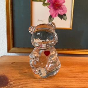 May include: A clear glass bear figurine with a red heart on its chest. The bear is sitting on a wooden surface, with a framed floral painting in the background. The bear is a decorative item.