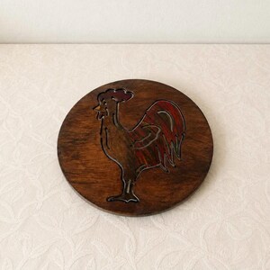Könnte beinhalten: Ein runder Untersetzer aus dunkelbraunem Holz mit einer eingravierten Hahn-Silhouette. Das Hahn-Motiv ist in tiefen Rot- und Brauntönen gehalten und zeigt die natürliche Maserung des Holzes auf der gesamten runden Oberfläche des Objekts.