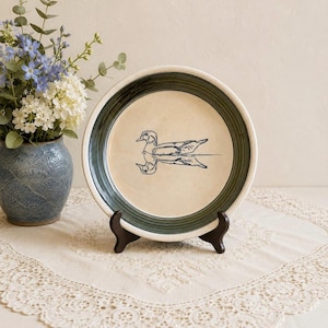 May include: A round ceramic plate featuring a dark blue line drawing of a duck swimming on water in the center. The plate has a wide, dark green rim and a cream-colored interior. It is displayed upright on a small black plastic stand.